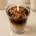 A lit candle in a ribbed clear glass cup, styled like iced coffee with dark and light layers, ice cubes, a small sprig of rosemary, and a visible flame. The candle is photographed on a warm cream surface with softly blurred white objects in the background.