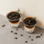 Two layered coffee-themed candles in clear glass cups, decorated with coffee beans, a sprig of rosemary, and dark chocolate pieces, set on a gray surface with scattered coffee beans against a cream wall.
