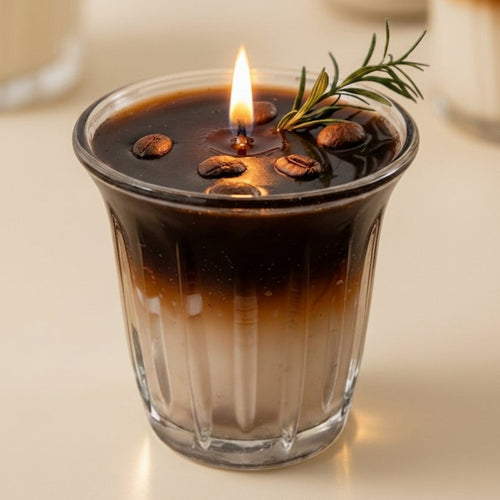 A lit candle in a ribbed clear glass cup, styled like an iced coffee drink with dark and light brown wax layers, coffee beans on top, and a green rosemary sprig garnish.