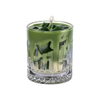 A green wax candle in a clear, faceted glass tumbler with a textured diamond-cut base, featuring a white wick at the top, set against a plain white background.
