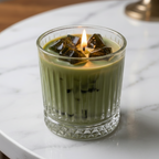 A lit candle in a ribbed clear glass cup, filled with light green wax and topped with dark green wax cubes, set on a white marble surface.