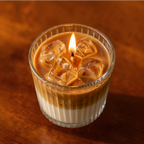A lit candle in a ribbed clear glass cup filled with layered coffee and milk textures, topped with ice cubes, on a warm wooden surface.